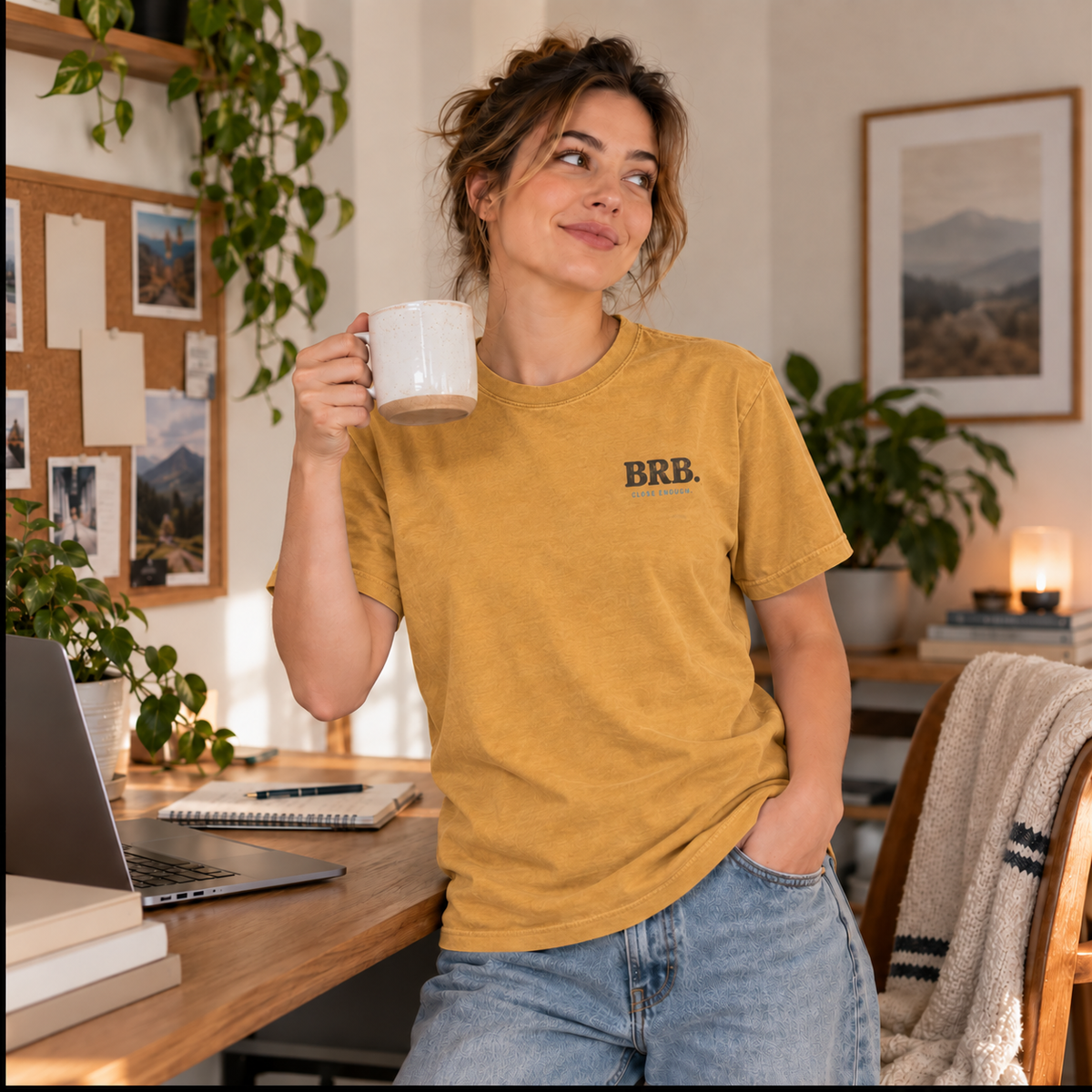 Female model wearing the Close Enough BE RIGHT BACK Tee in faded mustard during a coffee break in a warm home office